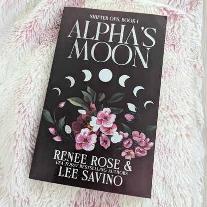 Image of the "Alpha's Moon" paperback with the alternate cover. It features pink flowers and the moon cycle. In white text is the title "Alpha's Moon." On the bottom of the cover is "Renee Rose & Lee Savino" and "USA Today Bestselling Authors"